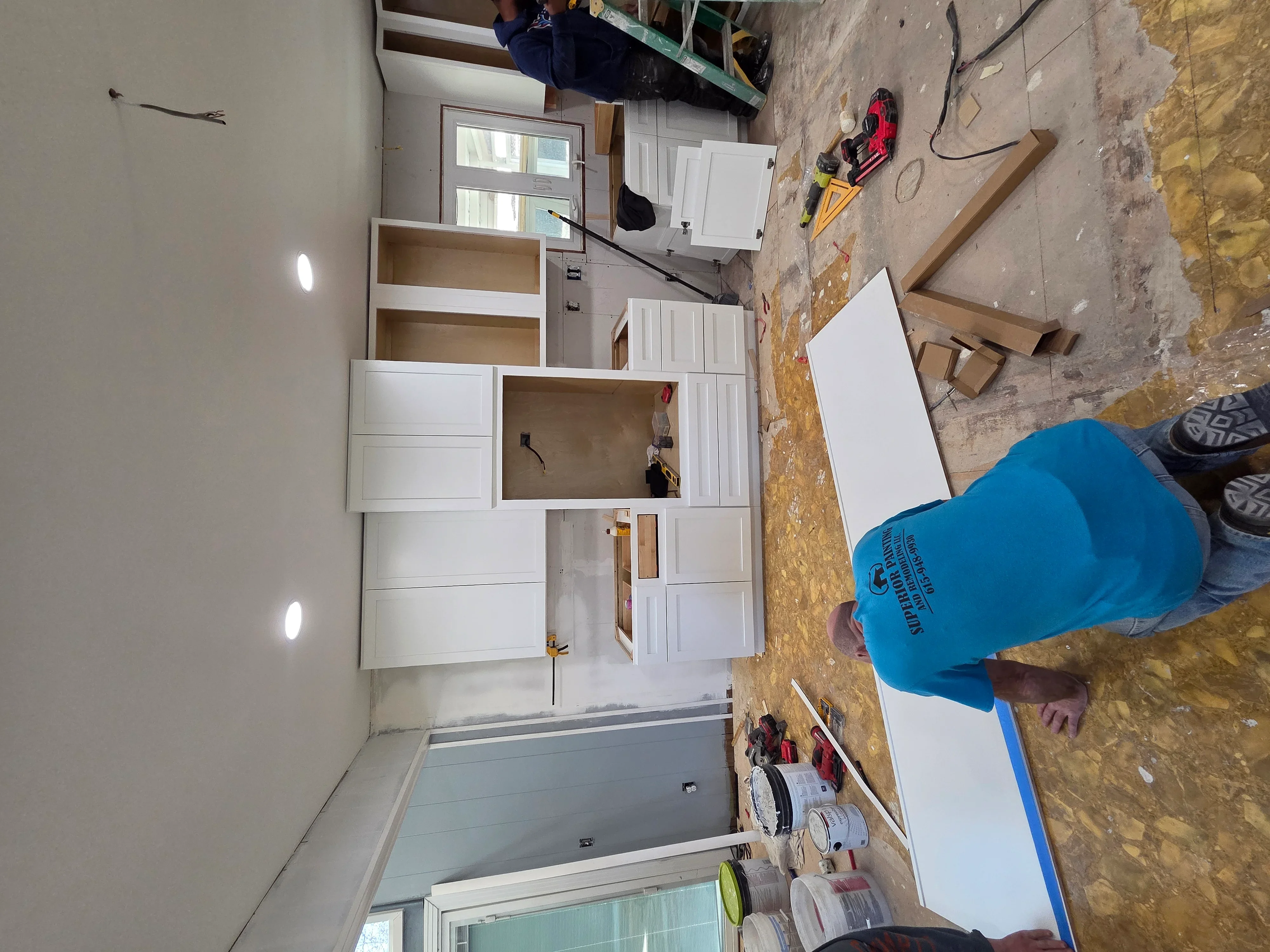 Kitchen remodel in progress showing white cabinetry installation with drywall finishing and recessed lighting